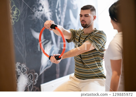Physical Therapy Session with Resistance Ring.A male patient performing arm exercises using a resistance ring under supervision during a rehabilitation session. 129885972