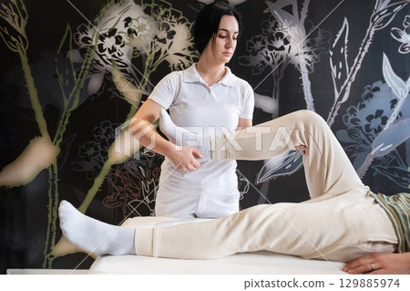 Physiotherapy Session with Blue Foam Roller.A therapist guiding a patient through leg exercises with the help of a blue foam roller in a therapy clinic. Physiotherapy Session with Blue Foam Roller.A therapist guiding a patient through leg exercises with the help of a blue foam roller in a therapy clinic. 129885974