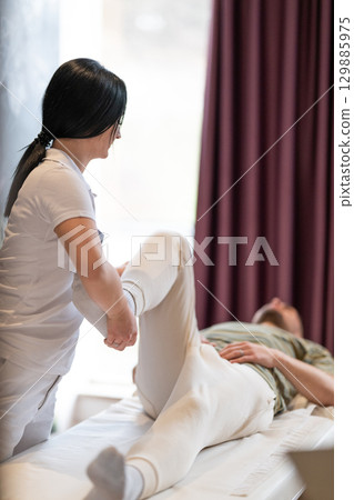 Physiotherapy Session with Blue Foam Roller.A therapist guiding a patient through leg exercises with the help of a blue foam roller in a therapy clinic. Physiotherapy Session with Blue Foam Roller.A therapist guiding a patient through leg exercises with the help of a blue foam roller in a therapy clinic. 129885975
