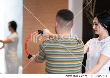 Physical Therapy Session with Resistance Ring.A male patient performing arm exercises using a resistance ring under supervision during a rehabilitation session. Physical Therapy Session with Resistance Ring.A male patient performing arm exercises using a resistance ring under supervision during a rehabilitation session. 129885981
