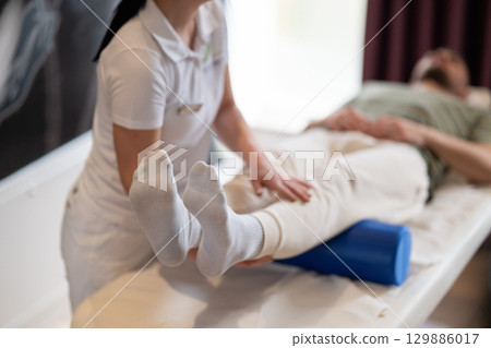 Physiotherapy Session with Blue Foam Roller.A therapist guiding a patient through leg exercises with the help of a blue foam roller in a therapy clinic. 129886017