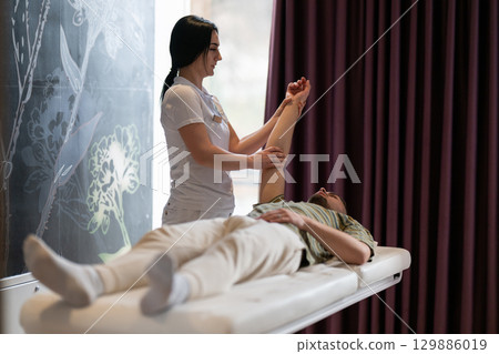 A female therapist performs a arm stretch technique on a male patient lying on a treatment bed. A female therapist performs a arm stretch technique on a male patient lying on a treatment bed. 129886019