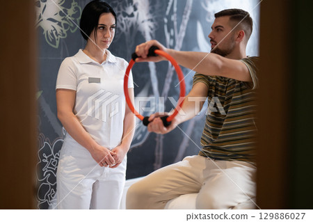 Physical Therapy Session with Resistance Ring.A male patient performing arm exercises using a resistance ring under supervision during a rehabilitation session. 129886027