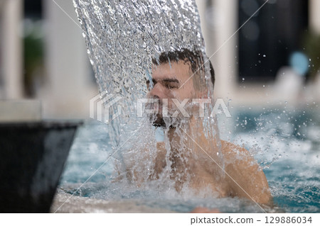 Man standing under a hydrotherapy waterfall in a spa pool, receiving water massage on his head and shoulders. Man standing under a hydrotherapy waterfall in a spa pool, receiving water massage on his head and shoulders. 129886034