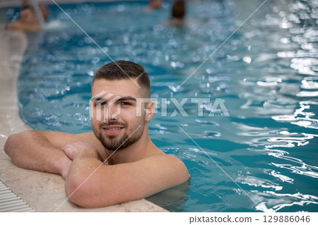 Man resting on the edge of a large indoor pool with a calm expression and relaxed posture. Man resting on the edge of a large indoor pool with a calm expression and relaxed posture. 129886046