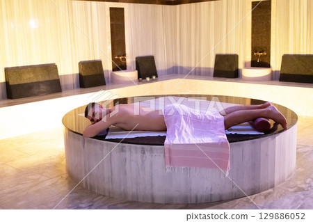 Man relaxing on a marble platform inside a Turkish bath with warm lighting. Man relaxing on a marble platform inside a Turkish bath with warm lighting. 129886052