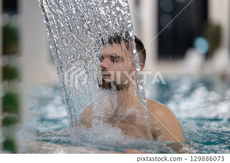 Man standing under a hydrotherapy waterfall in a spa pool, receiving water massage on his head and shoulders. 129886073