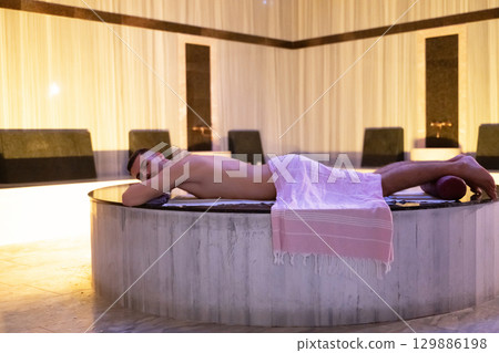 Man relaxing on a marble platform inside a Turkish bath with warm lighting. Man relaxing on a marble platform inside a Turkish bath with warm lighting. 129886198