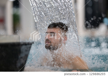 Man standing under a hydrotherapy waterfall in a spa pool, receiving water massage on his head and shoulders. 129886203
