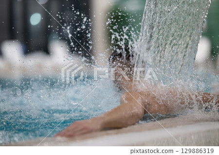 Man standing under a hydrotherapy waterfall in a spa pool, receiving water massage on his head and shoulders. Man standing under a hydrotherapy waterfall in a spa pool, receiving water massage on his head and shoulders. 129886319