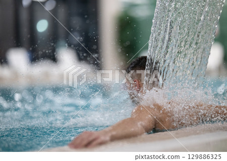 Man standing under a hydrotherapy waterfall in a spa pool, receiving water massage on his head and shoulders. Man standing under a hydrotherapy waterfall in a spa pool, receiving water massage on his head and shoulders. 129886325