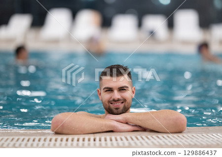 Man resting on the edge of a large indoor pool with a calm expression and relaxed posture. Man resting on the edge of a large indoor pool with a calm expression and relaxed posture. 129886437