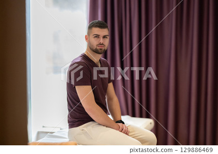A man sitting calmly on a treatment table, waiting for a physical therapy session in a clinic environment. 129886469