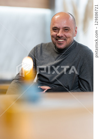 Smiling Man Sitting at Table.Smiling bald man in a turtleneck sweater sitting at a table with a candle in foreground. Smiling Man Sitting at Table.Smiling bald man in a turtleneck sweater sitting at a table with a candle in foreground. 129886721