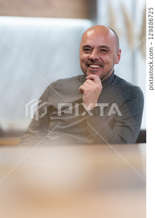 Smiling Man Sitting at Table.Smiling bald man in a turtleneck sweater sitting at a table with a candle in foreground. 129886725