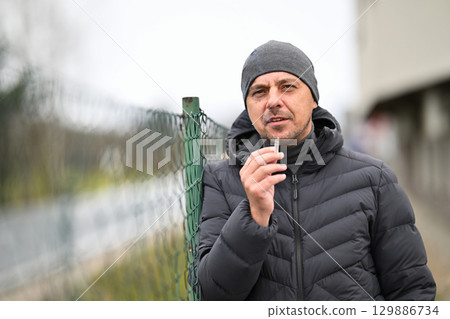 Man Smoking Outside.Man in winter jacket and beanie smoking a cigarette near a chain link fence outdoors. 129886734