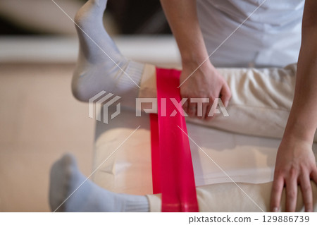Leg Therapy with Red Resistance Band.Close up view of a physiotherapist applying a red resistance band around a patient's ankles during a leg therapy session. 129886739