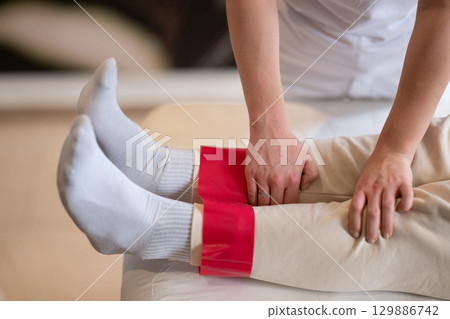 Leg Therapy with Red Resistance Band.Close up view of a physiotherapist applying a red resistance band around a patient's ankles during a leg therapy session. 129886742