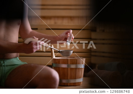 Essential Oils in Sauna. Close up of a mans hand holding essential oils inside a traditional wooden sauna with bucket and ladle. Essential Oils in Sauna. Close up of a mans hand holding essential oils inside a traditional wooden sauna with bucket and ladle. 129886748
