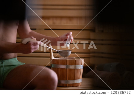 Essential Oils in Sauna. Close up of a mans hand holding essential oils inside a traditional wooden sauna with bucket and ladle. 129886752