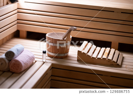 Sauna Bucket and Towels Close Up.Detailed close up of sauna bucket with ladle and essential oils next to rolled towels on wooden bench inside sauna. Sauna Bucket and Towels Close Up.Detailed close up of sauna bucket with ladle and essential oils next to rolled towels on wooden bench inside sauna. 129886755