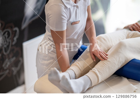 Physiotherapy Session with Blue Foam Roller.A therapist guiding a patient through leg exercises with the help of a blue foam roller in a therapy clinic. 129886756