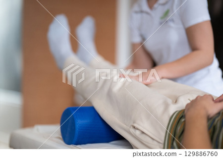 Physiotherapy Session with Blue Foam Roller.A therapist guiding a patient through leg exercises with the help of a blue foam roller in a therapy clinic. Physiotherapy Session with Blue Foam Roller.A therapist guiding a patient through leg exercises with the help of a blue foam roller in a therapy clinic. 129886760