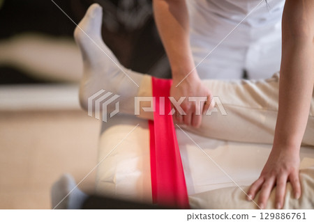Leg Therapy with Red Resistance Band.Close up view of a physiotherapist applying a red resistance band around a patient's ankles during a leg therapy session. Leg Therapy with Red Resistance Band.Close up view of a physiotherapist applying a red resistance band around a patient's ankles during a leg therapy session. 129886761
