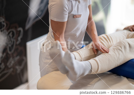 Physiotherapy Session with Blue Foam Roller.A therapist guiding a patient through leg exercises with the help of a blue foam roller in a therapy clinic. Physiotherapy Session with Blue Foam Roller.A therapist guiding a patient through leg exercises with the help of a blue foam roller in a therapy clinic. 129886766