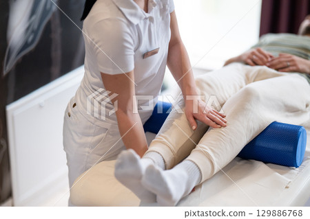 Physiotherapy Session with Blue Foam Roller.A therapist guiding a patient through leg exercises with the help of a blue foam roller in a therapy clinic. Physiotherapy Session with Blue Foam Roller.A therapist guiding a patient through leg exercises with the help of a blue foam roller in a therapy clinic. 129886768