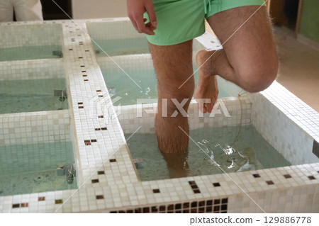 Man Stepping in Spa Water Walkway.Close up of a mans legs as he steps into tiled spa therapy pools for a hydrotherapy session. Man Stepping in Spa Water Walkway.Close up of a mans legs as he steps into tiled spa therapy pools for a hydrotherapy session. 129886778