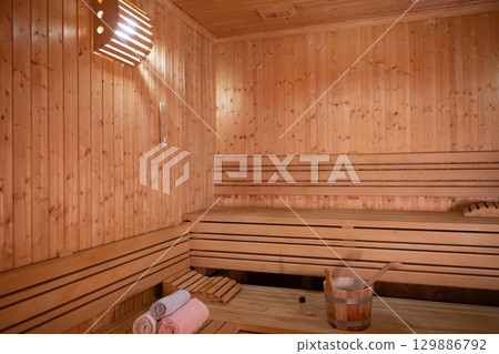 Empty Wooden Sauna Interior.Wide view of a warm and empty wooden sauna with rolled towels and wooden benches prepared for relaxation. Empty Wooden Sauna Interior.Wide view of a warm and empty wooden sauna with rolled towels and wooden benches prepared for relaxation. 129886792