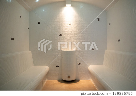Modern Steam Room Interior.Clean and minimalistic steam room with tiled walls and benches, featuring soft lighting and a central water fixture. 129886795