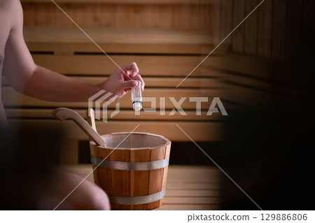 Essential Oils in Sauna. Close up of a mans hand holding essential oils inside a traditional wooden sauna with bucket and ladle. Essential Oils in Sauna. Close up of a mans hand holding essential oils inside a traditional wooden sauna with bucket and ladle. 129886806