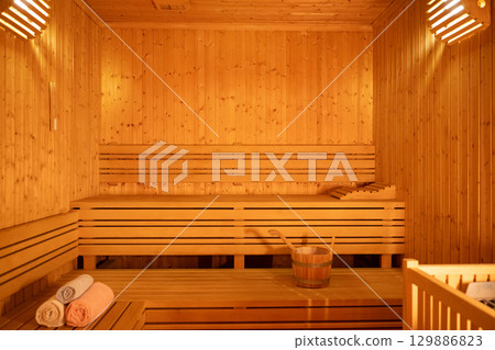 Empty Wooden Sauna Interior.Wide view of a warm and empty wooden sauna with rolled towels and wooden benches prepared for relaxation. 129886823