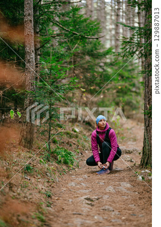 Woman Tying Shoe in Forest Trail.Female hiker crouching to tie her running shoe on a forest path surrounded by pine trees, dressed in sporty winter clothing. Woman Tying Shoe in Forest Trail.Female hiker crouching to tie her running shoe on a forest path surrounded by pine trees, dressed in sporty winter clothing. 129887013