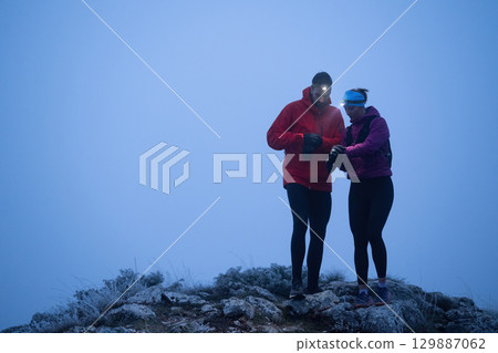 Checking GPS on the Summit.Two hikers consult their GPS device at the summit of a mountain during early morning with headlamps on, surrounded by mist. 129887062