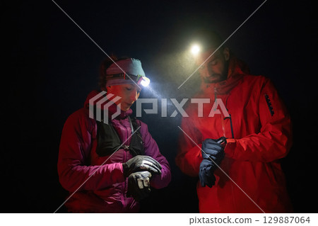Checking Coordinates During Night Hike.Couple night hikers examine a device under headlamp light, emphasizing navigation and teamwork under low light conditions. Checking Coordinates During Night Hike.Couple night hikers examine a device under headlamp light, emphasizing navigation and teamwork under low light conditions. 129887064