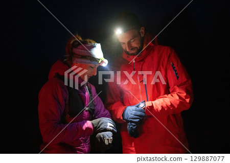 Checking Coordinates During Night Hike.Couple night hikers examine a device under headlamp light, emphasizing navigation and teamwork under low light conditions. Checking Coordinates During Night Hike.Couple night hikers examine a device under headlamp light, emphasizing navigation and teamwork under low light conditions. 129887077