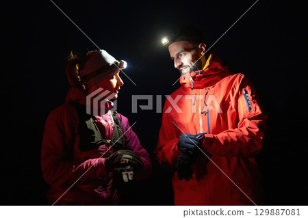 Checking Coordinates During Night Hike.Couple night hikers examine a device under headlamp light, emphasizing navigation and teamwork under low light conditions. 129887081