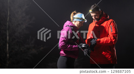 Checking Coordinates During Night Hike.Couple night hikers examine a device under headlamp light, emphasizing navigation and teamwork under low light conditions. Checking Coordinates During Night Hike.Couple night hikers examine a device under headlamp light, emphasizing navigation and teamwork under low light conditions. 129887105