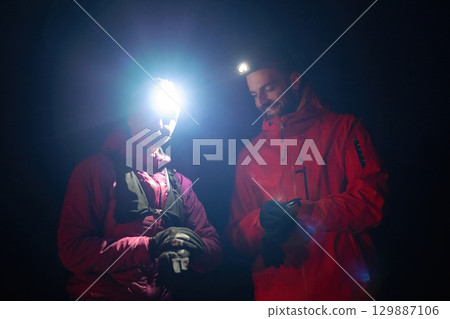 Checking Coordinates During Night Hike.Couple night hikers examine a device under headlamp light, emphasizing navigation and teamwork under low light conditions. 129887106