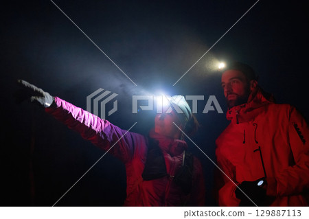 A woman points into the distance while discussing directions with her hiking partner under the beam of headlamps. 129887113