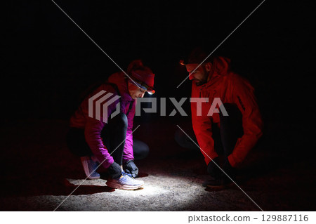 Night Trekking Footwear Check.Two trekkers inspect their shoes using headlamps during a night hike, emphasizing safety and preparation in the dark. 129887116