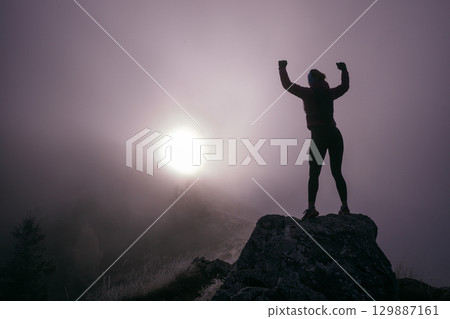 Woman Celebrating at Mountain Summit.Woman celebrating on rocky mountaintop with arms raised in victory surrounded by fog. Woman Celebrating at Mountain Summit.Woman celebrating on rocky mountaintop with arms raised in victory surrounded by fog. 129887161