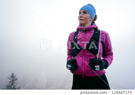 Confident female hiker in a pink jacket standing still on a misty mountain summit.Female Hiker Standing on Misty Peak 129887174