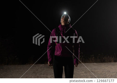 Woman Jogging at Night with Headlamp.Woman jogging at night with headlamp on, captured in dark outdoor environment. 129887177