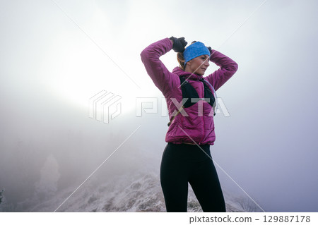 Active female hiker in pink jacket stretching arms on a foggy mountain peak during morning light. 129887178