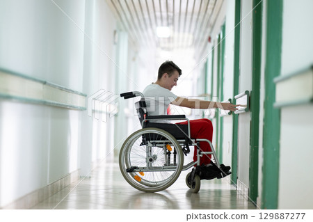 Young teenage boy smiling while sitting in a wheelchair in a hospital hallway. Rehabilitation and positivity concept. Young teenage boy smiling while sitting in a wheelchair in a hospital hallway. Rehabilitation and positivity concept. 129887277
