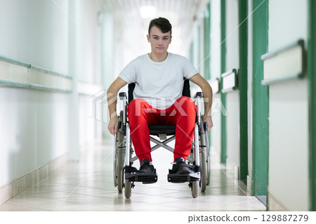 Young teenage boy smiling while sitting in a wheelchair in a hospital hallway. Rehabilitation and positivity concept. 129887279